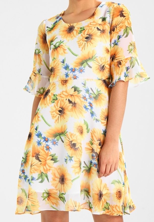 Femme portant une robe blanche avec de grands motifs de tournesol jaunes et de petites fleurs bleues, dotée de manches évasées trois-quarts et d'un col rond.