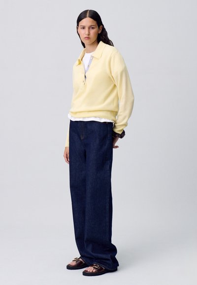 Suéter amarillo de manga larga con cuello, superpuesto a una camisa blanca. Jeans de pierna ancha azul oscuro y sandalias marrones con acentos metálicos.