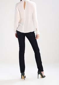 Blusa blanca de manga larga con abertura en la espalda, combinada con jeans de mezclilla oscuros y zapatos de tacón verdes. La blusa cuenta con puños fruncidos.