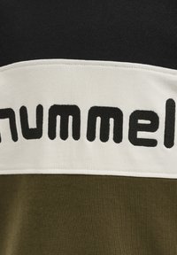 Olivově zelená tkanina s hladkým texturem obsahuje široký bílý pruh na vrchu, na kterém je výrazně napsáno slovo "hummel" černým písmem.