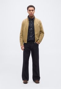 Άνδρας με μπεζ μπουφάν με φερμουάρ πάνω από σκοτεινό πουκάμισο με κουμπιά, φαρδιά σκούρα τζιν και καφέ παπούτσια, που στέκεται μπροστά σε απλό φωτεινό φόντο.