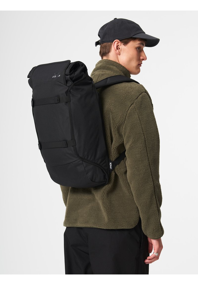 Black roll-top backpack with adjustable straps, textured fabric, and minimalistic design. Visible brand tag on the side.