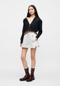 Νεαρή γυναίκα που φορεί μαύρο φούτερ με φερμουάρ, καφέ μπλούζα, ανοιχτό γκρι μίνι φούστα και σκούρα καφέ μπότες μέχρι τη μέση της μηριαίας περιοχής, στέκεται με το ένα χέρι στην τσέπη.