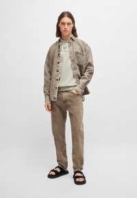 Rjava teksturirana denim jakna s žepom, svetlo zelena polo majica in usklajene hlače. Črne sandale dopolnjujejo obleko.