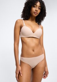 Γυναίκα με σγουρά μαλλιά που φοράει σουτιέν σε γυμνό χρώμα και ταιριαστό εσώρουχο, στέκεται μπροστά σε απλό ανοιχτό φόντο.