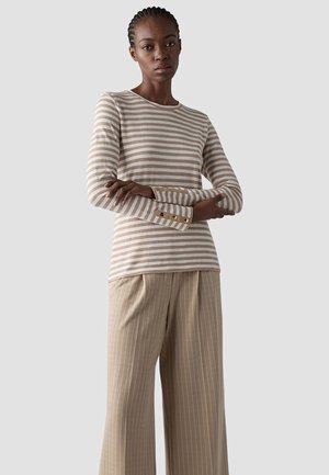 Woman with braided hair wears beige and white striped long-sleeve top and beige pinstripe wide-leg trousers, arms crossed, neutral background.