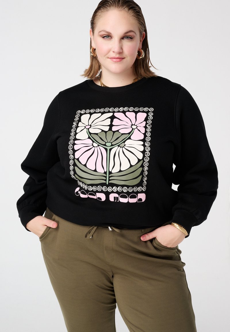 Femme aux cheveux courts portant des boucles d'oreilles créoles en or, un sweat-shirt noir avec un motif floral, et un pantalon vert olive les mains dans les poches.