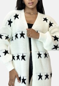 Cárdigan de punto blanco con patrón de estrellas negras; tiene apertura frontal, mangas largas y puños acanalados. Textura suave y ajuste relajado.