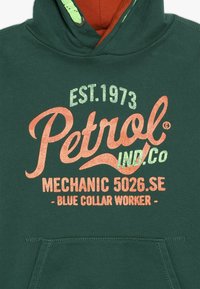 Zelená mikina s klokaní kapsou, se slovy v oranžové a zelené barvě. Design obsahuje "Petrol IND. Co." a "Modrý límeček".