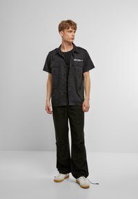 Camicia nera a maniche corte con bottoni con stampa "SECURITY", dotata di due tasche frontali e una texture liscia. Abbinata a pantaloni neri e sneakers.