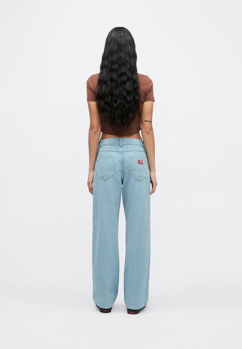 Vrouw met lang golvend haar, gekleed in een bruin cropped top en lichtblauwe wijde jeans, staand met haar rug naar de camera.