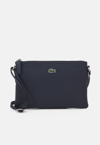 Pochette en tissu texturé bleu marine avec fermeture éclair et bracelet, ornée d'un petit logo brodé de crocodile sur le devant.