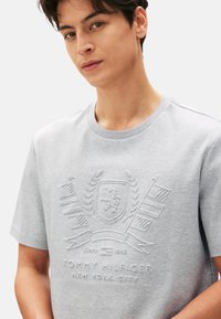 Camiseta de color gris claro hecha de tela suave, con un diseño de logo y emblema texturizado. Cuello redondo clásico y mangas cortas.