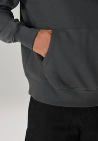 Felpa grigio scuro con tasca frontale, polsini a costine e finitura strutturata, abbinata a pantaloni neri.