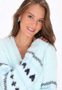 Ljusblå tröja med V-urringning, mjukt material och mönstrade ärmar som har svarta och blå designs. Modellen bär blå örhängen.