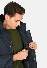 Giacca blu navy con zip, cappuccio e bottoni a pressione. Presenta una tasca interna con zip e un maglione lavorato a maglia verde oliva sotto.