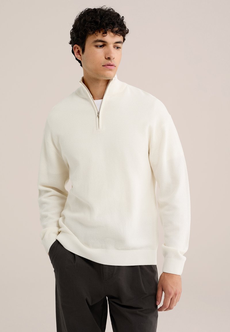 Jeune homme portant un pull blanc texturé à demi-fermeture éclair et un pantalon noir, debout avec une main dans la poche, regardant vers le bas sur le côté.