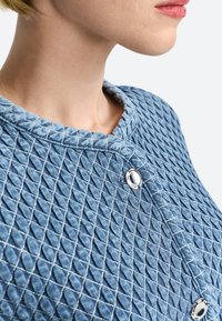 Giacca di jeans trapuntata in azzurro chiaro con motivo esagonale, caratterizzata da un colletto rotondo e dettagli in bottoni argentati. Tessuto testurizzato.