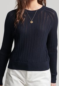 Granatowy sweter dzianinowy z prążkowaną strukturą, długimi rękawami, okrągłym dekoltem oraz dopasowanym wykończeniem. Naszyjnik z złotym wisiorkiem stanowi subtelny akcent.