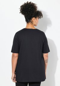 Černé tričko, volný střih, krátké rukávy, kulatý výstřih, hladká bavlněná tkanina. Model zobrazen zezadu, zdůrazňující výstřih a délku rukávů.