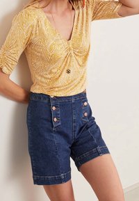 Žlutý vzorovaný top s krátkými rukávy, kombinovaný s tmavými džínovými šortkami s vysokým pasem, které mají dekorativní knoflíky a tapered střih.