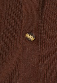 Brązowy sweter z prążkowanej dzianiny w zbliżeniu, z okrągłym guzikem z dwoma otworami, mającym ciemniejszą krawędź i jaśniejszy środek. Widoczne detale teksturowanej dzianiny.