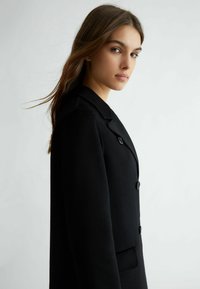 Cappotto in lana nera con colletto a punta, due bottoni frontali e tasca laterale. Texture liscia con una vestibilità sartoriale.