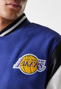 Μπλε μάλλινο varsity μπουφάν με μαύρες δερμάτινες λεπτομέρειες και ένα εμφανές κεντητό λογότυπο των Los Angeles Lakers σε κίτρινο και μωβ χρώμα.