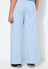 Světle modré široké kalhoty s pruhy a jemnou texturou, svislé bílé pruhy a volný střih. Viditelné černé platformové boty.