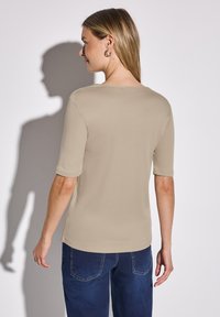 Beige košile s krátkým rukávem a kulatým výstřihem, hladká textura a volný střih, kombinovaná s tmavě modrými džínami.