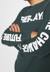 Person with folded arms wearing a dark long-sleeve shirt with white text "REPLAY" on chest and "FUTURE" and "CHANGE" on sleeves.