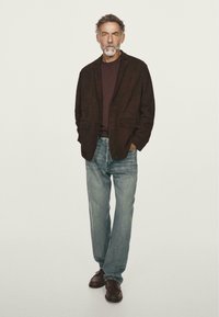 Blazer marrone scuro sopra una camicia bordeaux, abbinato a jeans azzurri chiari e scarpe marroni. Il blazer ha due tasche frontali e una vestibilità comoda.