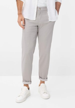 Man die lichtgrijze opgerolde broek, witte sneakers en een wit, dichtgeknoopt overhemd draagt met handen in de zakken.