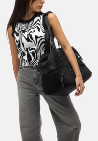 Schwarze Reisetasche mit Netzpaneel, ausgestattet mit mehreren Reißverschlusstaschen und dekorativen Metallteilen. Das Model trägt ein schwarz-weiß gemustertes Oberteil und graue Hosen.