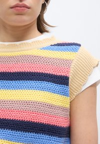 Gros plan sur une personne portant un gilet tricoté à rayures multicolores par-dessus une chemise blanche, montrant l'épaule et une partie du visage.