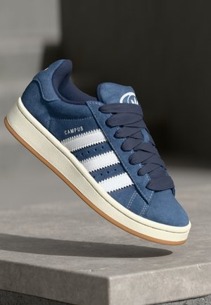 Niebieskie tenisówki Adidas Campus z białymi paskami, ciemnoniebieskimi sznurowadłami i gumową podeszwą unoszące się nad kamienną powierzchnią.