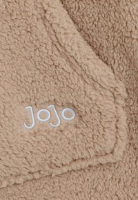 Béžová fleece látka s textúrovaným povrchom, s vyšitým logom "Jojo" v bielej farbe. Dizajn obsahuje viditeľné švy.