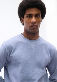 Νέος άνδρας με σκούρα σγουρά μαλλιά, φορώντας ένα ανοιχτό γαλάζιο πουκάμισο με υφή και μακριά μανίκια, κοιτώντας κατευθείαν την κάμερα με ουδέτερη έκφραση.