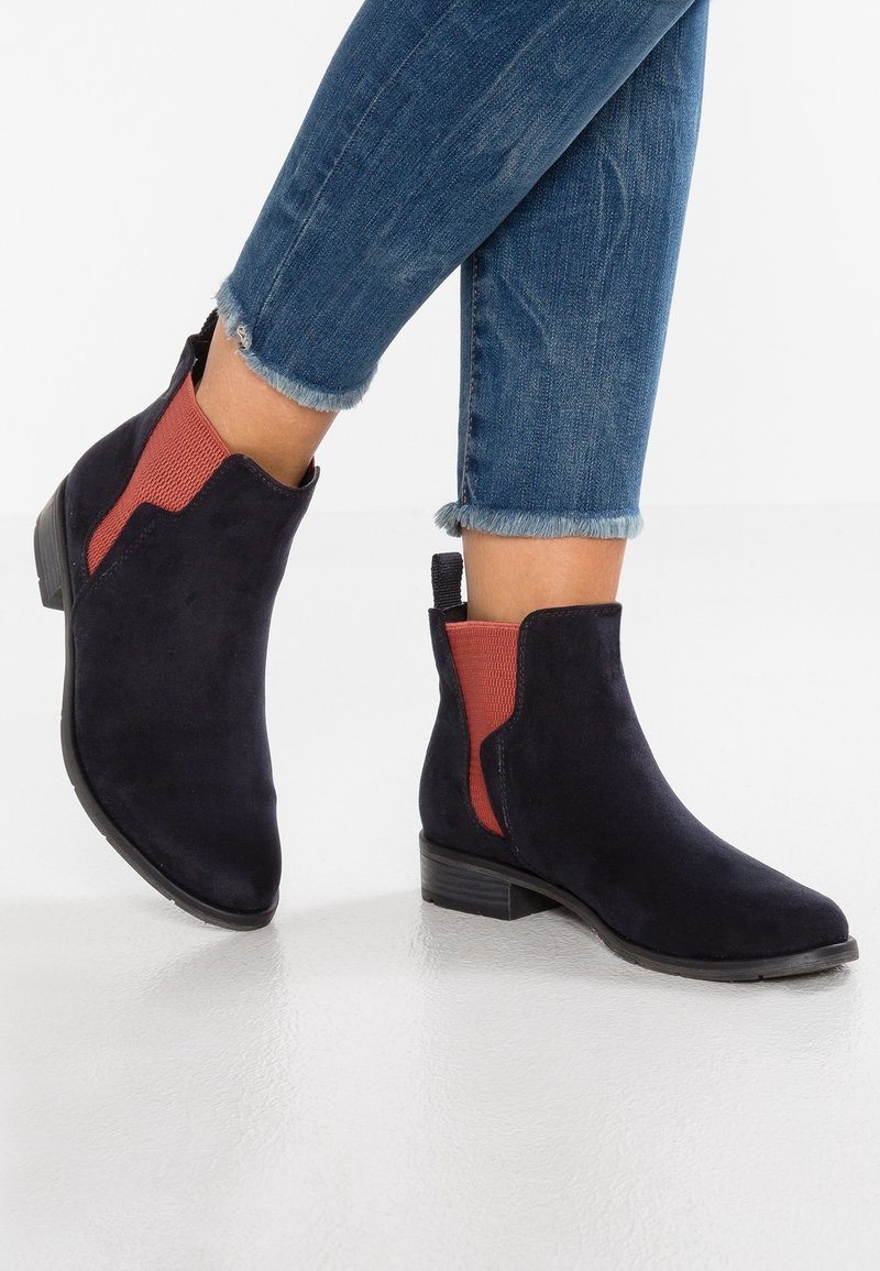 Bottines en daim noir avec panneaux latéraux élastiques de couleur rouille, talon plat et semelle en caoutchouc texturée. Jean avec ourlet effiloché visible.