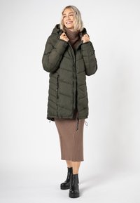 Lächelnde Frau in olivgrünem Pufferjacke, braunem Rollkragenkleid und schwarzen Chelsea-Stiefeletten, vor einem weißen Hintergrund stehend.