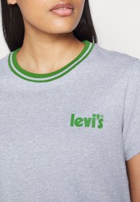 Femme portant un t-shirt Levi's gris clair avec un col rayé vert et un logo Levi's vert sur la poitrine, montrant le cou et le haut du torse.