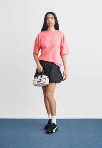 Γυναίκα που στέκεται φορώντας ροζ T-shirt Puma, μαύρο αθλητικό σορτς, μαύρα αθλητικά παπούτσια Puma, λευκές κάλτσες και κρατώντας μια λευκή τσάντα Puma.