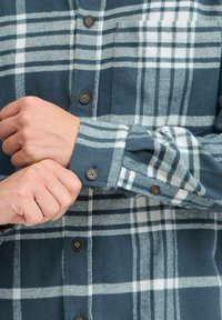 Green and white plaid flannel shirt with brown buttons and a chest pocket; close-up of sleeves being adjusted. Soft texture and warm material.