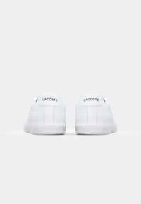 Sneakers albe din piele cu o textură netedă, având un design low-top, vârf rotund și logo-ul verde "LACOSTE" pe călcâi.