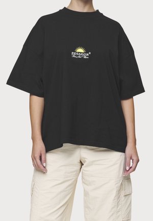 Osoba nosząca luźną czarną koszulkę z małym żółtym słońcem i białym logo "PEGADOR", połączoną z jasnobeżowymi spodniami typu cargo.