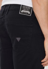 Vaqueros de mezclilla negra con un primer plano del bolsillo trasero que presenta un parche triangular de logo "GUESS" en rojo y negro con costuras blancas.