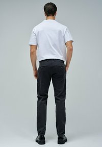Uomo che indossa una camicia bianca a maniche corte, pantaloni scuri e scarpe nere, in piedi con le spalle rivolte verso un semplice sfondo grigio chiaro.
