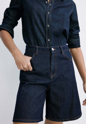 Person iført en mørkeblå denim skjorte med knapper, som er stukket ned i matchende højtaljede denimshorts, med den ene hånd i lommen.