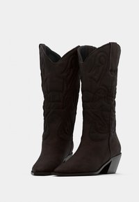 Σκούρα καφέ καουμπόικες μπότες μήκους έως τη μέση της γάμπας με κεντητά σχέδια σπειροειδούς μορφής και γωνιώδεις ξύλινες φτέρνες σε λευκό φόντο.