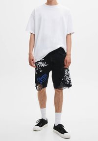T-shirt blanc oversize, shorts en denim noir déchirés avec des graphiques et du texte bleus. Porté avec des chaussettes blanches et des baskets noires.